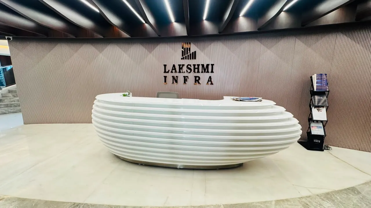 A ribbed white reception desk in front of a fluted timber wall carrying the Lakshmi Infra name, under a dark timber blade ceiling